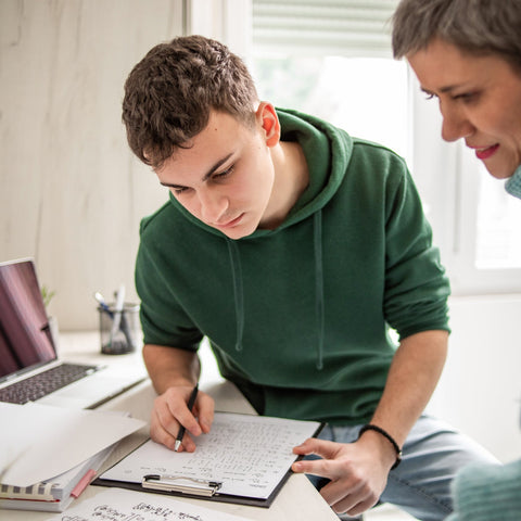 A teenage boy and a woman working together on a project with a laptop and papers in a bright room.