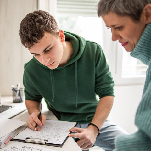 A teenage boy and a woman working together on a project with a laptop and papers in a bright room.