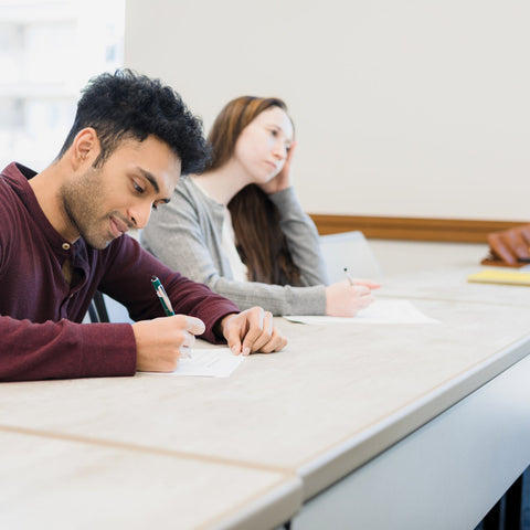 Two students sitting at a table in a classroom, taking notes.