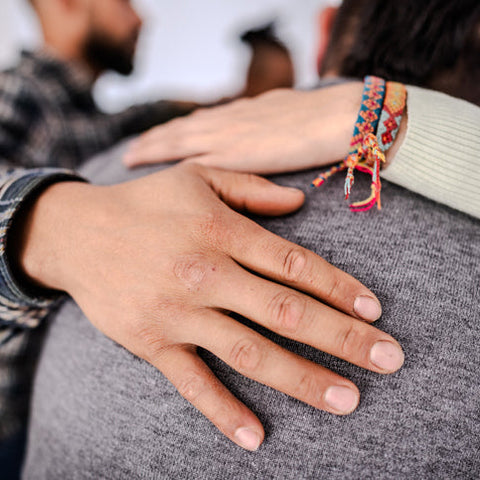 Person wearing a colorful bracelet on a gray sweater with another person's hand on their shoulder.