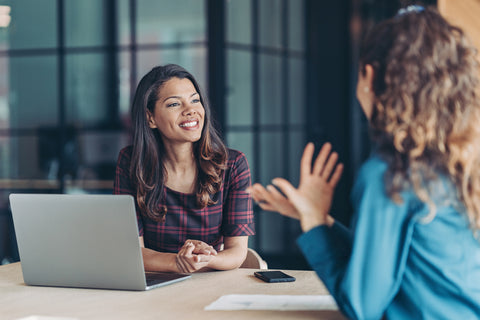 Two women sitting at a desk with a laptop, engaged in a conversation.