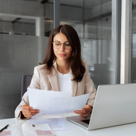 Woman in an office setting holding papers and using a laptop.