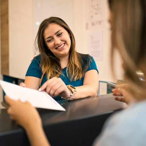 Friendly staff member at a front desk smiling while handing paperwork to a student, demonstrating a welcoming customer service interaction.