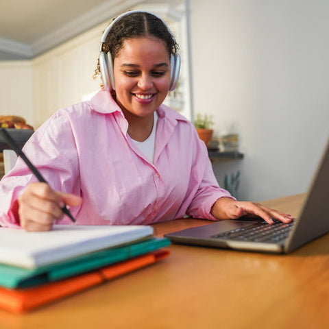 Woman in pink shirt with headphones using laptop at a table with notebooks
