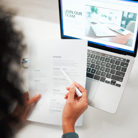 Person reviewing a printed resume with a laptop screen showing a 'Join Our Team' job posting in the background.