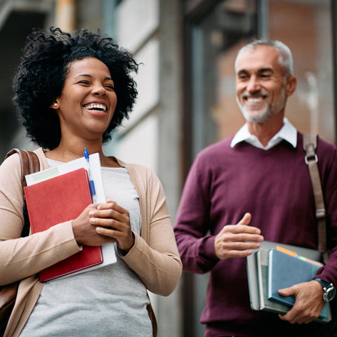 Two people walking outdoors, one holding books and a bag, the other with a shoulder bag.