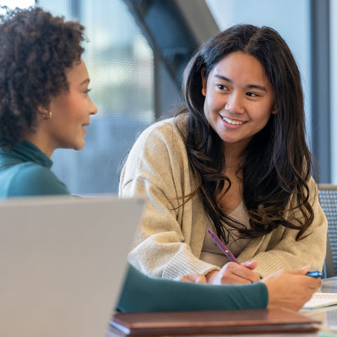 Two women sitting at a table with a laptop, engaged in a conversation.