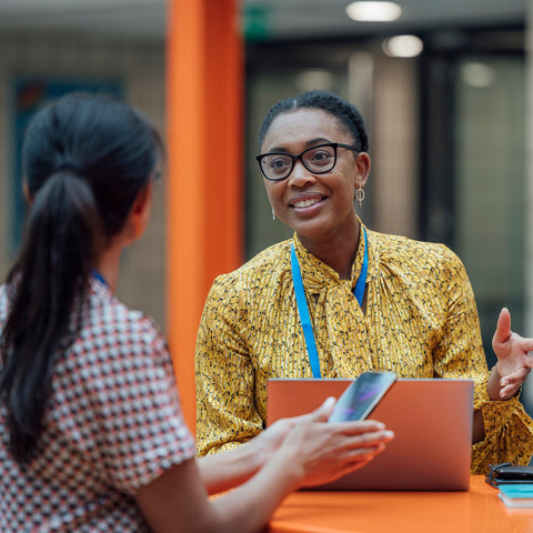 A staff member with a lanyard talks with a student at a table, laptop and notebooks nearby.