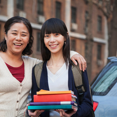 A mother and her college-aged daughter smile together outside campus housing, daughter holding textbooks and wearing a backpack.