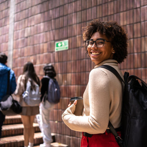 Woman with a backpack smiling in front of a brick wall with people in the background