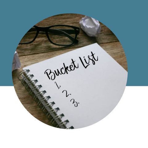 Notebook with 'Bucket List' on a wooden surface with glasses and a crumpled piece of paper.