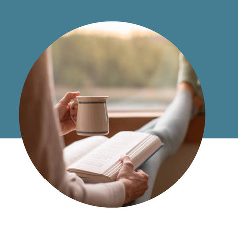 Person reading a book with a mug, against a scenic window view.