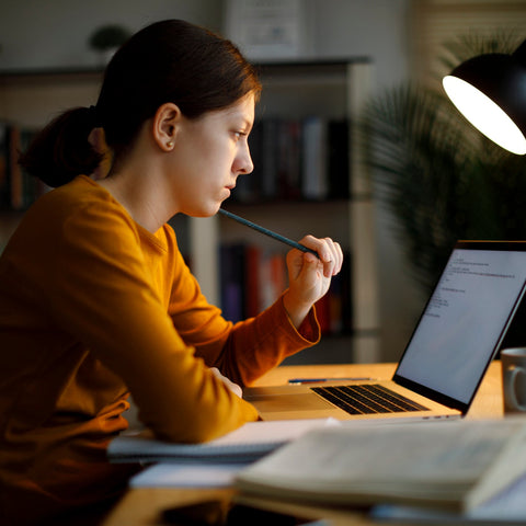 Student studying at a desk at night, thoughtfully reviewing content on a laptop while holding a pencil, focused and engaged.
