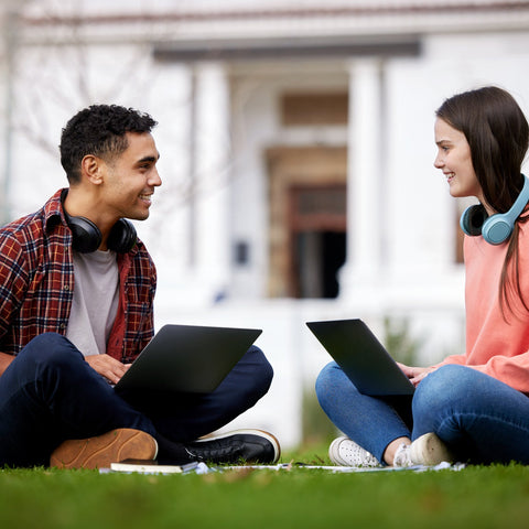 Two students sitting on grass with laptops in front of a building