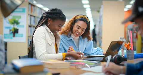Two women studying together in a library with books and a laptop.