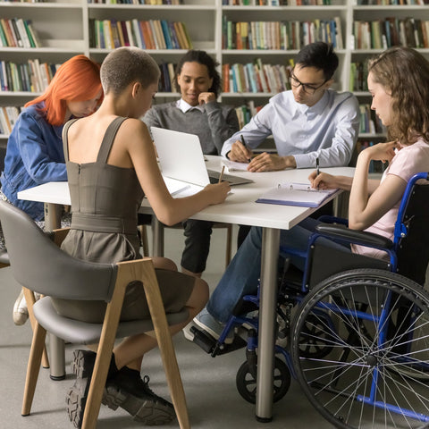 Group of people, including a person in a wheelchair, sitting around a table in a library.