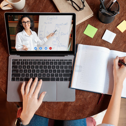 Person attending a virtual meeting on a laptop with notes and a coffee cup on a wooden table.