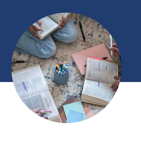 An overhead view of someone surrounded by open books, colorful pens, and notebooks while studying on the floor.