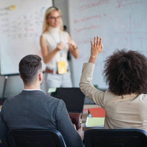 A participant raises their hand during a professional training session led by a presenter at a whiteboard.