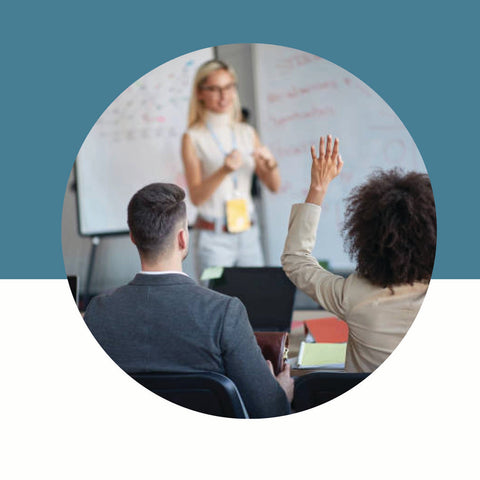 Two people in a classroom setting with a teacher at the front, one raising a hand.