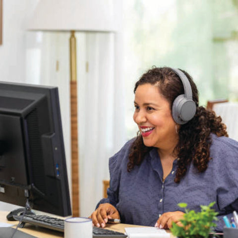 A smiling woman wearing headphones works at a desktop computer in a bright home office setting.
