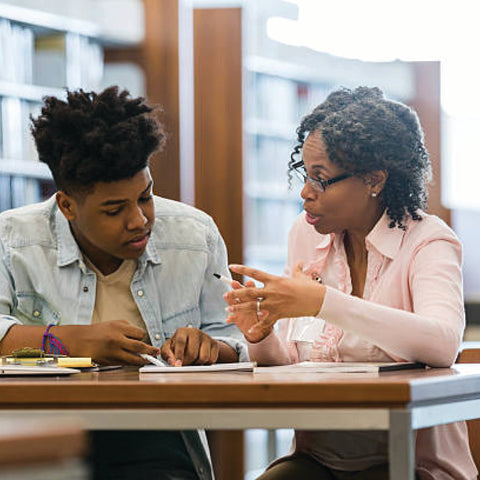A tutor and student sit together at a library table, reviewing notes in an engaged one-on-one session.