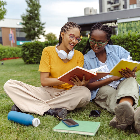 Two people sitting on grass outdoors, reading books.