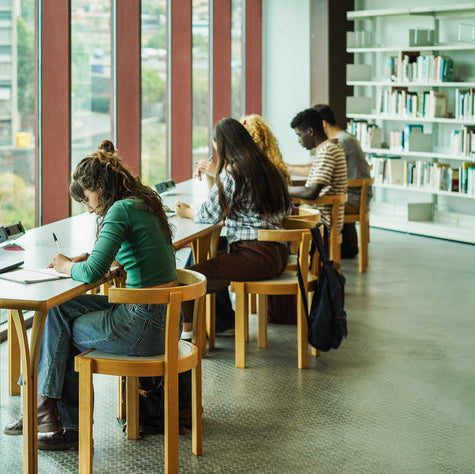 Students studying in a library with large windows and bookshelves.