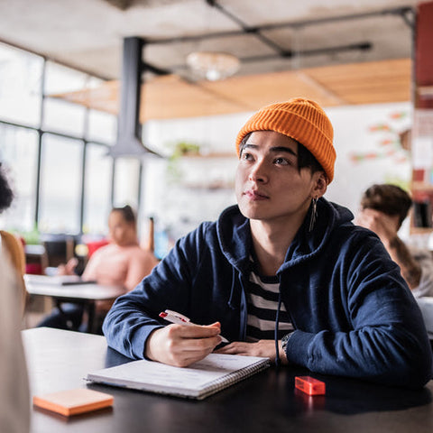 Person wearing an orange beanie and blue hoodie sitting at a table in a classroom setting.