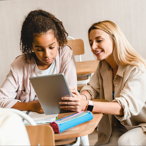 Teacher kneeling beside student desk helping young girl with tablet in classroom