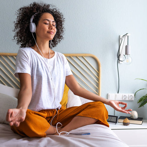 Woman meditating on a bed with headphones on, in a bedroom setting.