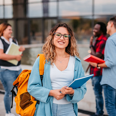 A young woman with glasses and a yellow backpack holds notebooks and smiles at the camera while three other people talk in the background outside a building.