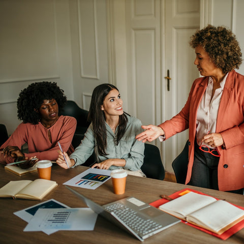 Group of women in a meeting around a table with laptops and documents.