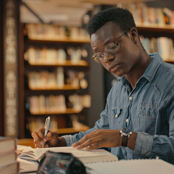 Man studying in a library with books and a laptop on a table.