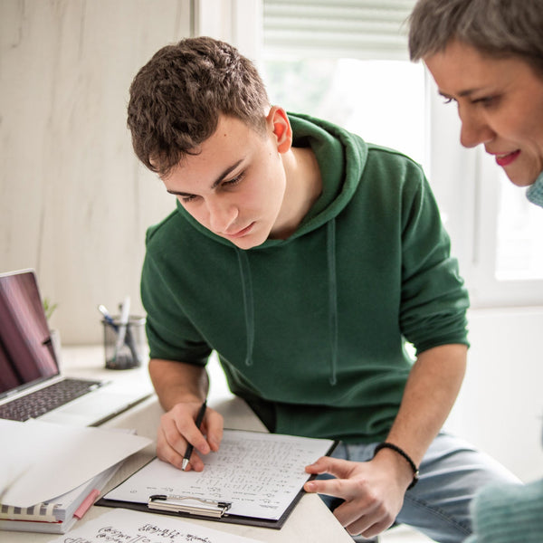 A teenage boy and a woman working together on a project with a laptop and papers in a bright room.