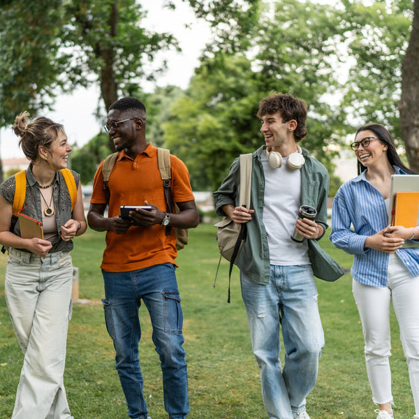 Four college students walking and laughing together on a campus lawn, carrying backpacks, notebooks, and a laptop.