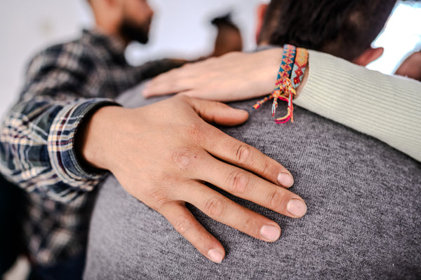Person wearing a colorful bracelet on a gray sweater with another person's hand on their shoulder.