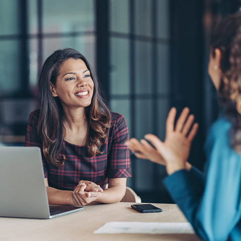 Two women sitting at a desk with a laptop, engaged in a conversation.