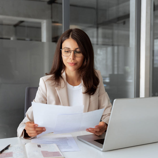 Woman in an office setting holding papers and using a laptop.