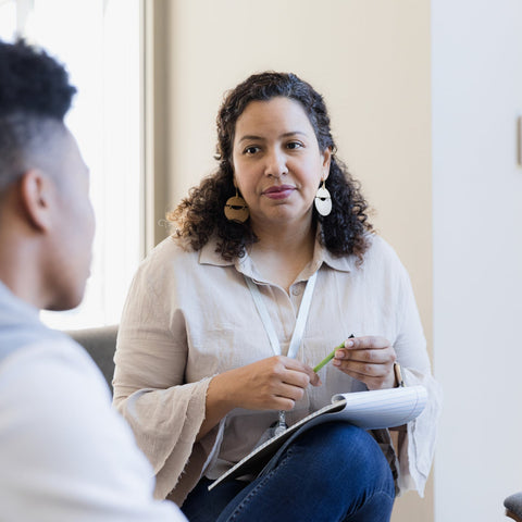 Woman in a professional setting holding a pen and clipboard, engaged in conversation with another person.