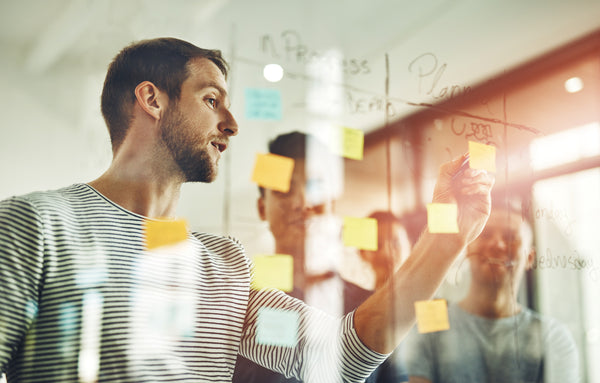 Two people working together on a glass board with sticky notes in an office setting.
