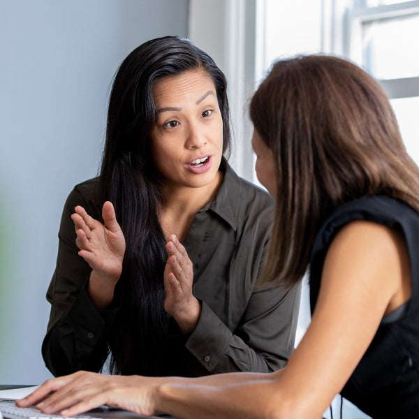Two women in an office reviewing information together at a laptop, one gesturing while speaking