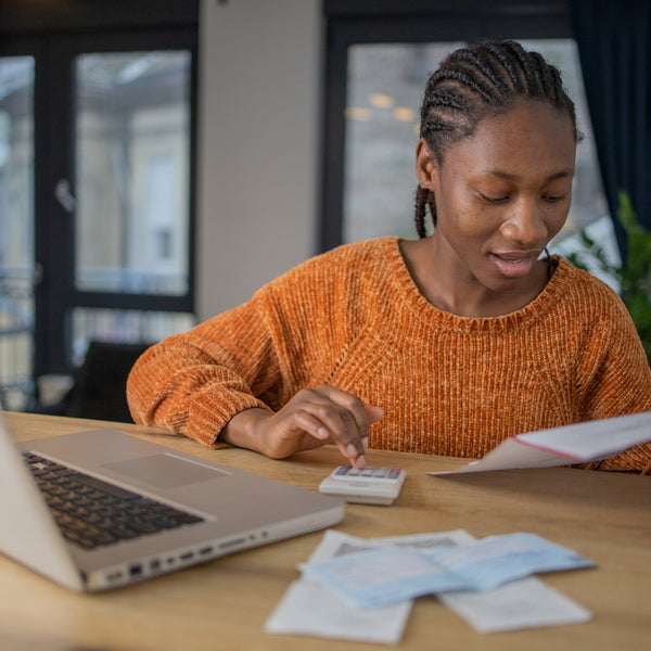 Woman in an orange sweater sitting at a desk with a laptop, calculator, and papers.