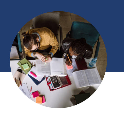 An overhead view of two students studying together at a desk covered in open books, a calculator, and colorful sticky notes.