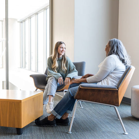 Student and advisor sitting in comfortable chairs having a supportive one-on-one conversation, representing coaching, reflection, and building resilience.