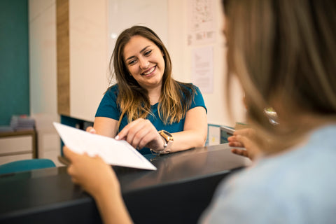 Friendly staff member at a front desk smiling while handing paperwork to a student, demonstrating a welcoming customer service interaction.