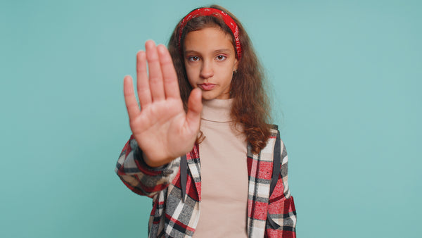 Teenage girl facing camera with palm raised, signaling "stop," against a teal background.

