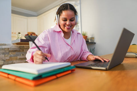 Woman in pink shirt with headphones using laptop at a table with notebooks