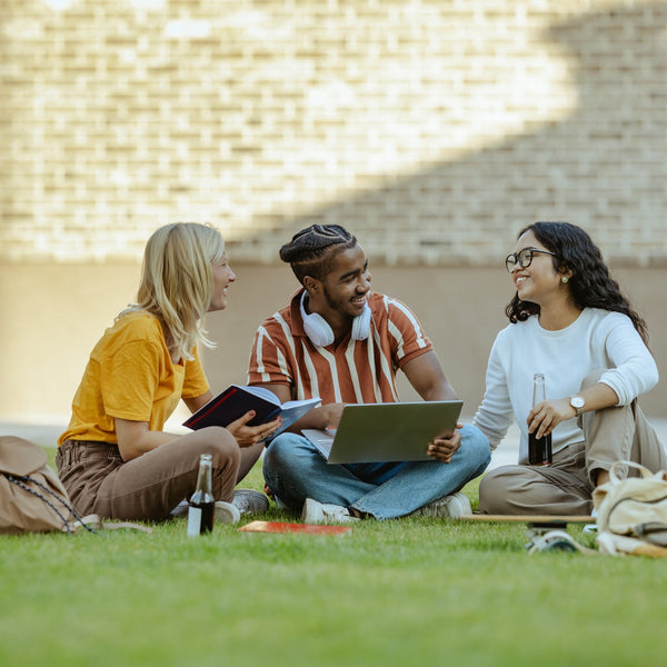 Three students sitting on grass with books and a laptop