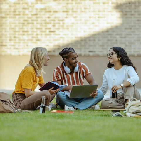 Three students sitting on grass with books and a laptop